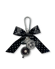 A black and silver handcrafted bag charm featuring a polka dot bow and various charms, attached to a stainless steel carabiner hook.